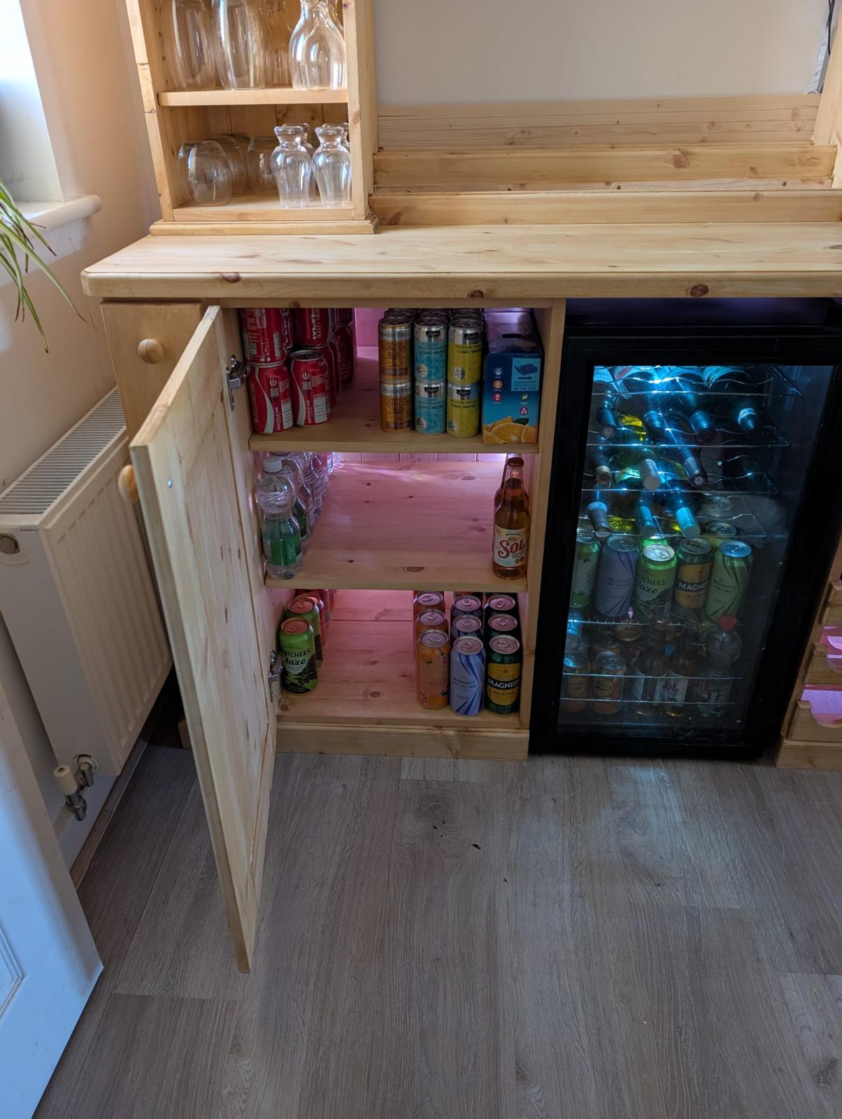 Open lower cupboard stocked with mixers, beers and tonics, with the glassware shelves above.